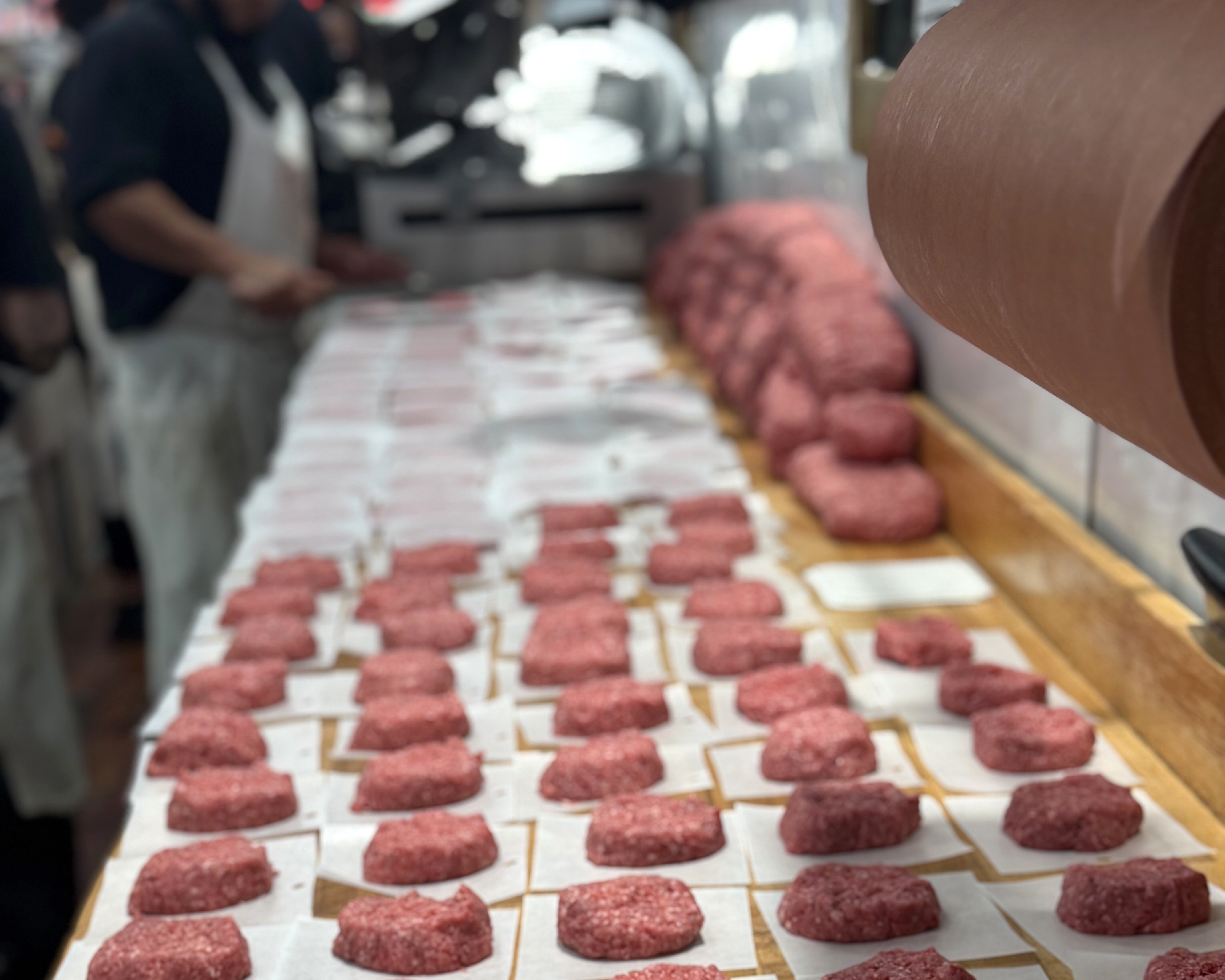 A butcher shaping fresh-ground burger patties by hand at the counter, rows of formed patties laid out beside him